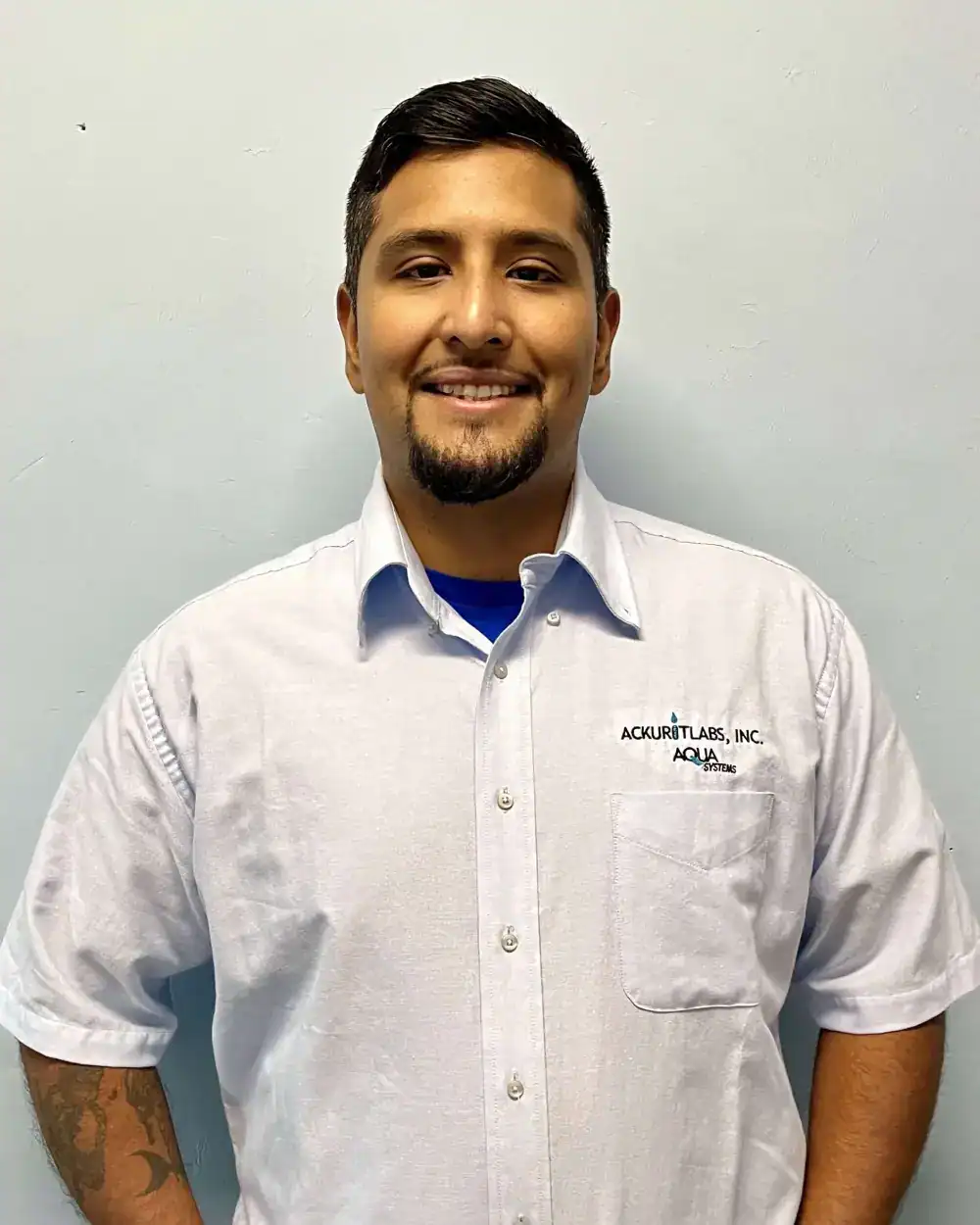 Male technician in white shirt with Ackuritlabs and Aqua Systems logo, smiling, against plain wall.