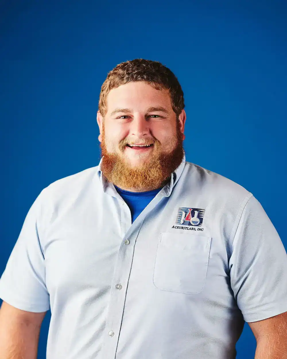 Smiling technician in light blue uniform with Ackuritlabs logo against blue background.