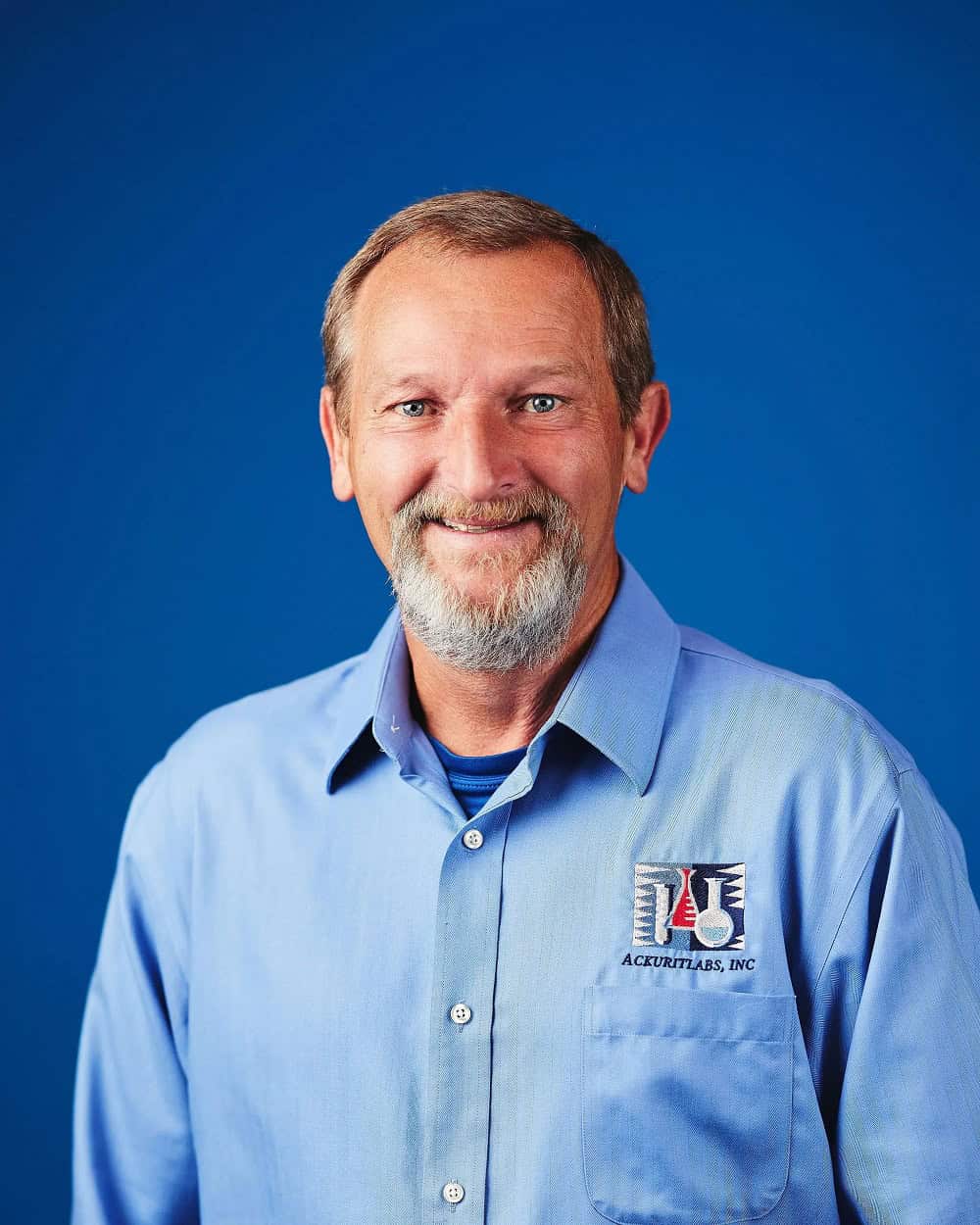 Portrait of a smiling man in a blue work shirt with Ackuritlabs logo against a blue background.
