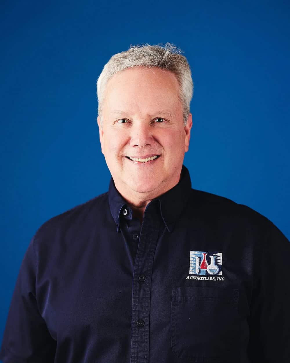 Portrait of a smiling man in a black shirt with Ackuritlabs logo against a blue background.