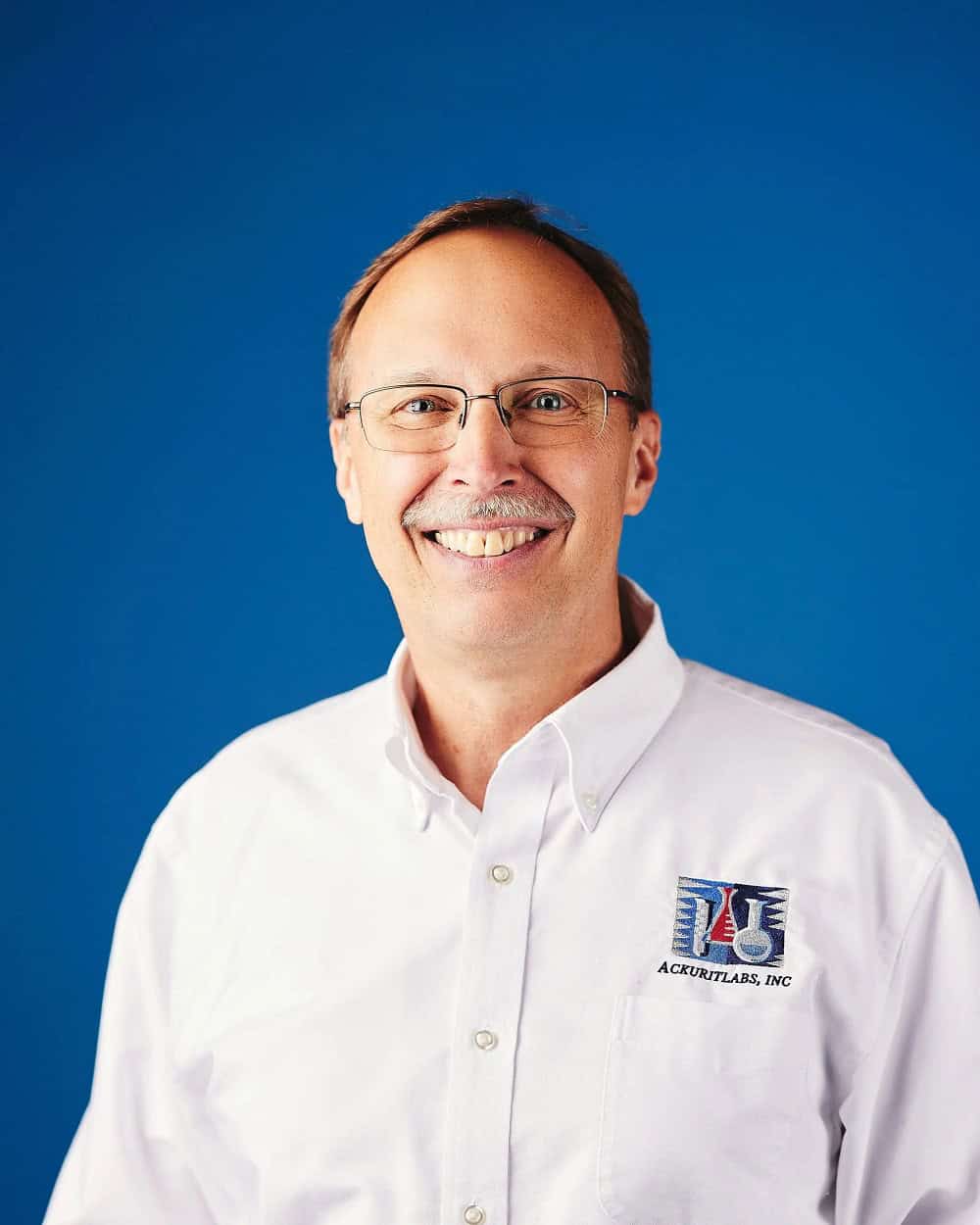 Smiling man in white shirt with Ackuritlabs logo against blue background.