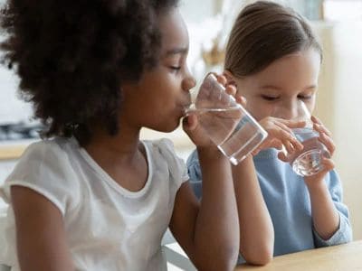 Two young girls drinking water from clear glasses at a table.