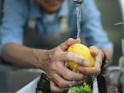 Person washing a lemon under a running faucet in a kitchen sink.