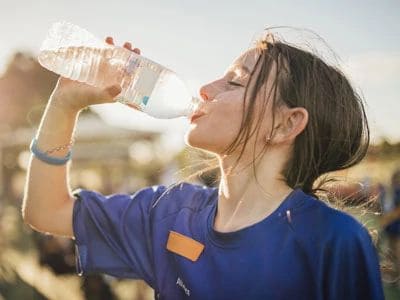 Young woman drinking water outdoors, wearing a blue shirt, with sunlight in the background.