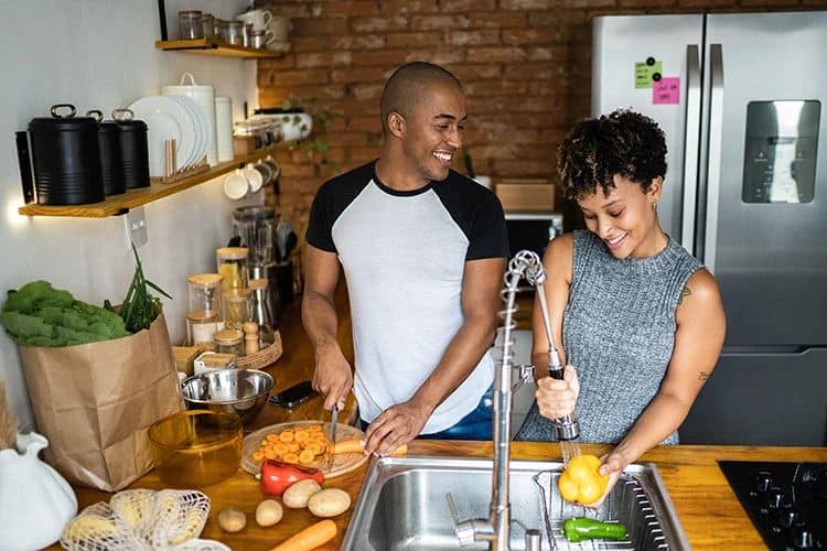 Two people preparing food in a modern kitchen with a brick wall and stainless steel appliances.