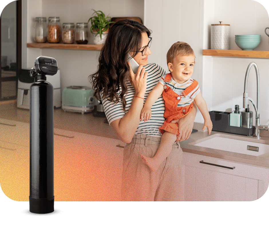 Woman with glasses holding a child in a modern kitchen, talking on the phone, with a water testing device nearby.