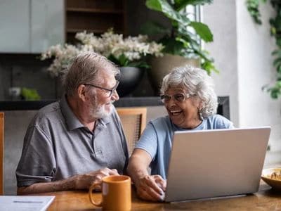 Elderly couple using a laptop at home, smiling and talking.