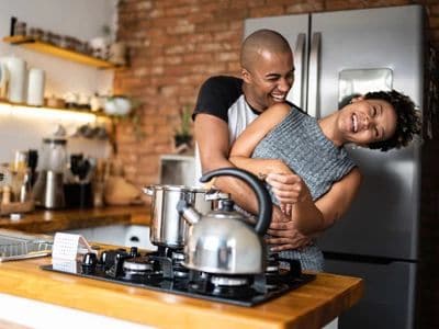 A man and woman laughing in a kitchen with a stove and kettle.