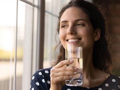 Woman drinking water, smiling, near a window in a bright room.